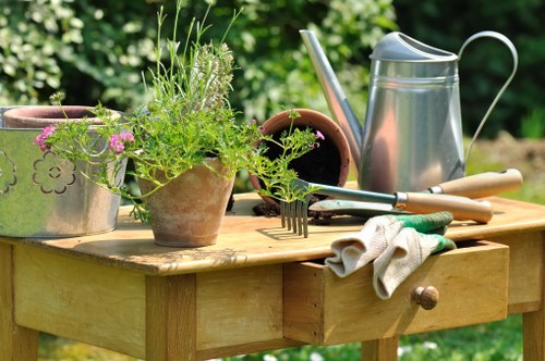 A wooden outdoor table in a garden setting is shown, topped with various gardening tools and accessories. There are potted plants with green foliage and small purple flowers, a metal watering can, and a few gardening hand tools such as a rake and trowel, some of which are resting on a pair of gardening gloves. The background features lush green trees and shrubs, indicating a well-maintained garden area, possibly in Seven Kings. The scene suggests active gardening or landscaping work, with natural sunlight illuminating the workspace, and the overall environment appears tidy and inviting for outdoor garden maintenance or planting activities. This setup exemplifies typical garden care activities catered to local outdoor spaces, aligning with the services offered by Gardening Services Seven Kings.