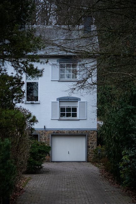 A winding cobblestone driveway leading to a red brick house with a tiled roof, partially obscured by lush greenery including mature trees, trimmed bushes, and climbing plants on the house walls. The d